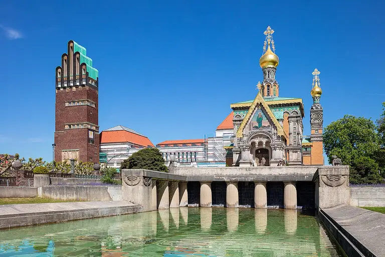 Der Hochzeitsturm und die Gebäude der Künstlerkolonie auf der Mathildenhöhe in Darmstadt, fotografiert im Querformat unter blauem Himmel.